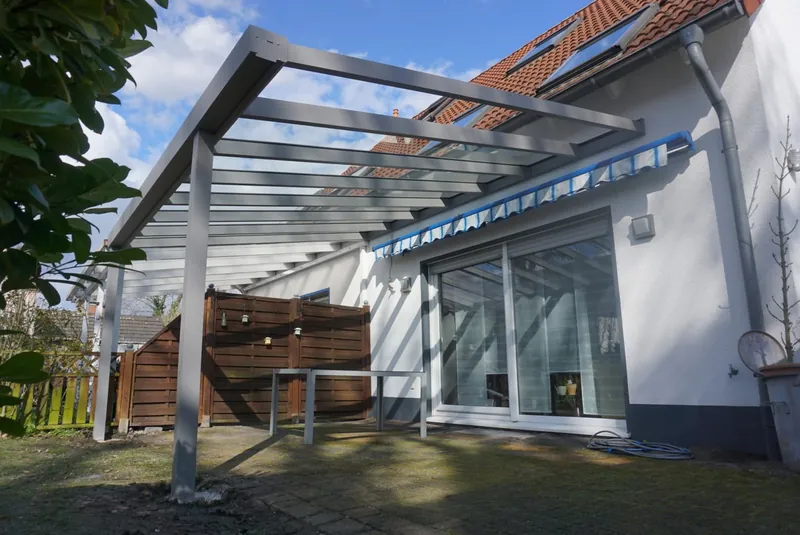 Szare aluminiowe zadaszenie ze szklanym dachem i markizą przy białym domu