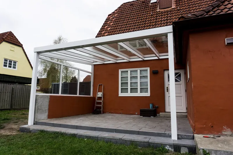 Białe zadaszenie tarasu ze szklanym dachem i szklaną ścianą boczną przy domu pod czerwoną dachówką